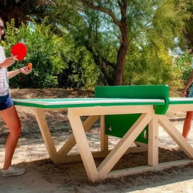Deux femmes jouent au ping-pong à l'extérieur au Camping Ensoya, sous un ciel bleu et des arbres verts.