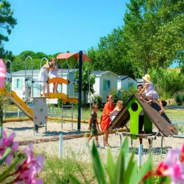 Enfants jouent sur un parc de jeux en bois au Camping Ensoya, entouré de fleurs roses et de cabanes.