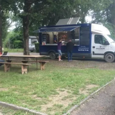 Camping Le Plantaurel : camion-restaurant 'SAUDADE' ouvert, clients et personne sur banc sous arbre.