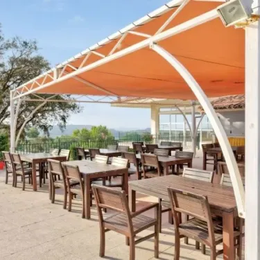 Terrasse avec tables et chaises sous une toile orange au Camping Le Parc des Chênes.