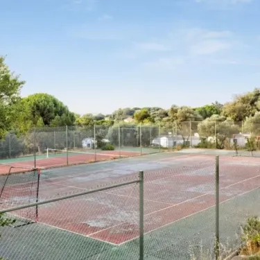 Court de tennis extérieur au Camping Le Parc des Chênes, entouré d'arbres et de clôtures.