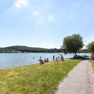Camping Yaloer : personnes se relaxant sur l'herbe près d'un lac, chemin de terre et arbres