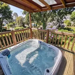 Jacuzzi sur une terrasse en bois sous un auvent, entouré de verdure et de haies.