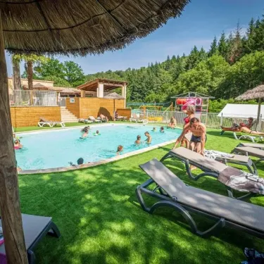 Piscine ovale du Camping Coeur d'Ardèche avec parasols en paille, chaises longues et enfants nageant.