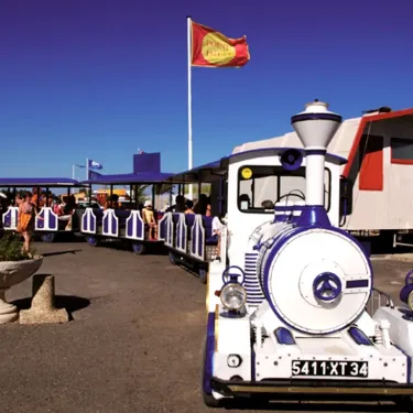 Train touristique blanc et bleu au Camping Le Castellas, sous un drapeau rouge.