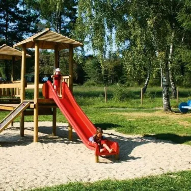 Enfant glissant sur une slide rouge dans un parc de jeux en bois au Camping du Lac de Miel.