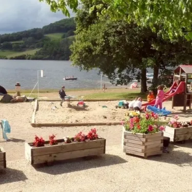 Enfants jouent au sable près d'un parc infantile avec un lac en arrière-plan à Camping La Romiguière.