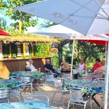 Terrasse extérieure du camping Le Bourdieu avec parasols, tables et chaises autour d'un bar tropical.