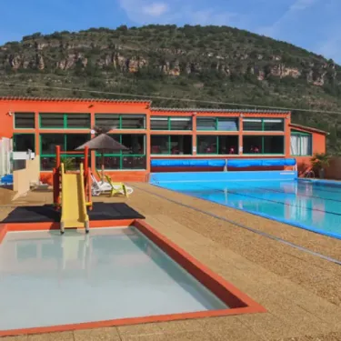 Camping La Cascade - piscine entourée de chaises longues et d'un bâtiment orange sur fond de montagne