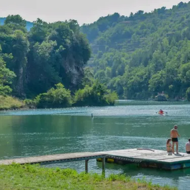 Camping La Cascade : ponton en bois au bord d'un lac avec des visiteurs et des kayaks.