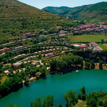 Vue aérienne d'un village niché près d'un lac turquoise, entouré de collines vertes et de routes serpentines.