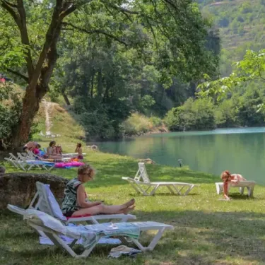 Camping La Cascade : personnes se relaxant sur des transats sous les arbres à côté d'un lac.