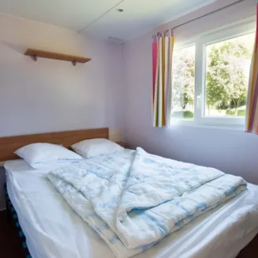 Chambre dans le Mobil Home Confort Tuffault, lit double, rideaux colorés, étagère murale.