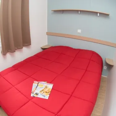 Chambre Mobil Home Eden Thouet avec lit rouge, étagères murales, rideaux marron.