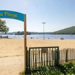 Entrée de la plage avec panneau 'Plage', clôture verte et lac en arrière-plan