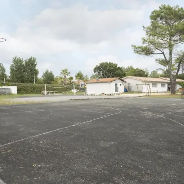 Terrain de basket-ball au Camping Paradis Albret, entouré d'arbres et de bâtiments blancs.