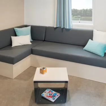 Mobil Home Smala 4 Chambres 8 Personnes - Camping La Touesse : canapé L en tissu gris avec coussins bleus et blancs, table basse en bois et métal, vue sur la fenêtre.