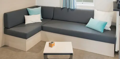 Mobil Home Smala 4 Chambres 8 Personnes - Camping La Touesse : canapé L en tissu gris avec coussins bleus et blancs, table basse en bois et métal, vue sur la fenêtre.