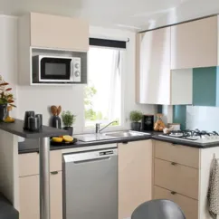 Mobil Home Smala 4 Chambres 8 Personnes - Camping La Touesse : cuisine moderne avec micro-ondes, lave-vaisselle, planche de cuisson, étagères et fenêtre.