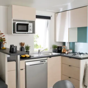 Mobil Home Smala 4 Chambres 8 Personnes - Camping La Touesse : cuisine moderne avec micro-ondes, lave-vaisselle, planche de cuisson, étagères et fenêtre.