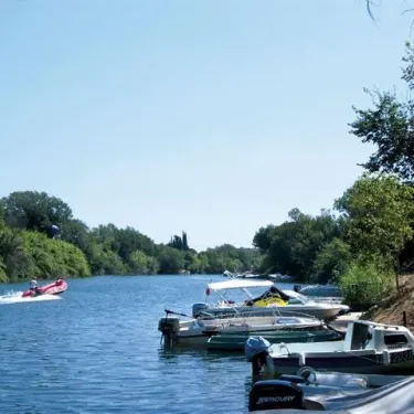 Camping La Barque : embarcation sur un fleuve avec des bateaux amarrés et une embarcation en cours de navigation.