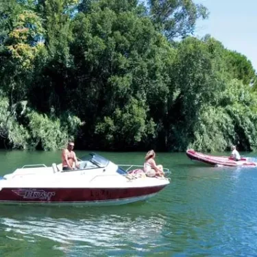 Un couple navigue en bateau motorisé sur une rivière entourée de forêt dense, Camping La Barque.