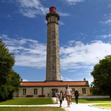 Phare historique sous un ciel bleu, avec des visiteurs marchant sur une allée verte près du Camping La Côte Sauvage.