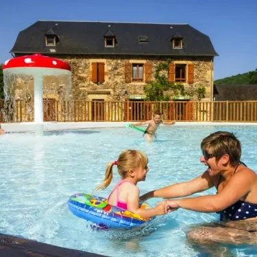 Une mère aide sa fille dans la piscine à Camping La Boissière, près d'un jouet flottant sous un abri en forme de champignon.