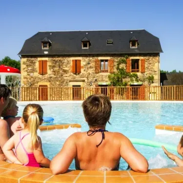 Une famille profite de la piscine à Camping La Boissière, avec une maison en pierre en arrière-plan.