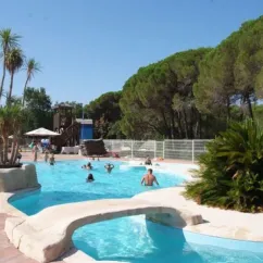 Piscine avec toboggan et palmiers au Camping Oasis Village, visiteurs se baignent sous un ciel bleu.