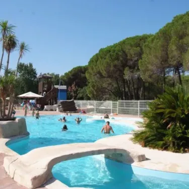Piscine avec toboggan et palmiers au Camping Oasis Village, visiteurs se baignent sous un ciel bleu.