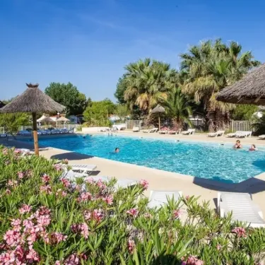 Piscine avec chaises longues et parasols en paille au Camping Beau Rivage, bordée de fleurs roses.