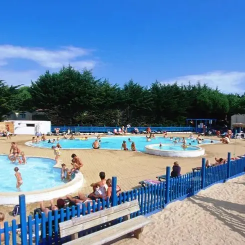 Piscine extérieure animée avec des enfants et adultes, entourée de chaises longues et de clôtures bleues.