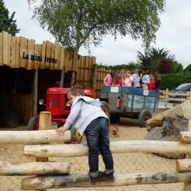 Enfant grimpe sur un fossé en bois à Camping La Grand Métairie, près d'un tracteur rouge et d'une clôture en bois.