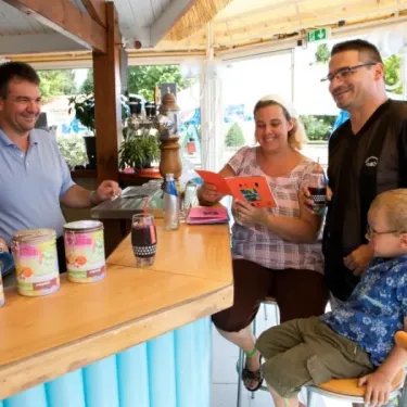 Famille au bar du Camping La Grand Métairie, dégustant des boissons et des produits locaux sous une pergola en paille.