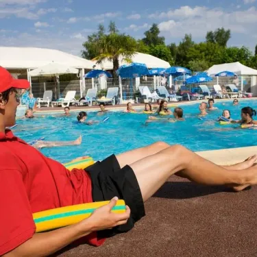 Moniteur de piscine en tenue rouge près d'une grande piscine entourée d'ombrelles et de chaises longues.