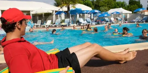 Moniteur de piscine en tenue rouge près d'une grande piscine entourée d'ombrelles et de chaises longues.