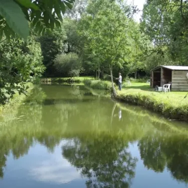 Un pêcheur sur un étang entouré de forêt, avec une cabane en bois, Camping Etangs Fleuris