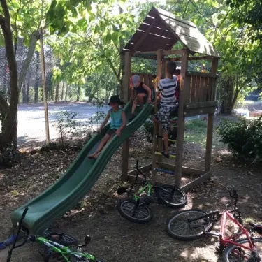 Enfants jouent sur une aire de jeu avec toboggan vert et vélos autour, dans le parc du Camping Val Roma Park.