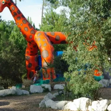 Camping Des Pins : girafe gonflable colorée dans un parc avec arbres et rochers.