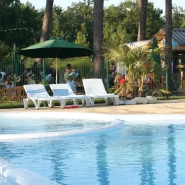 Camping Des Pins : chaises longues blanches près de la piscine, parasol vert, pins en arrière-plan
