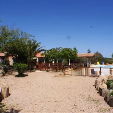 Allée de gravier bordée de rochers et de végétation au Camping La Trinité, avec une piscine en fond.