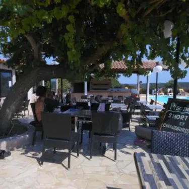 Camping La Trinité : terrasse ombrée sous arbre avec tables et chaises, panneau indiquant piscine short et chaussures.