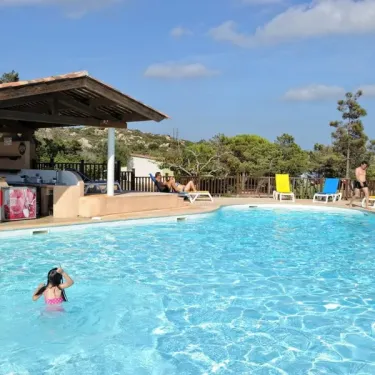 Une enfant nage dans la piscine du Camping La Trinité, avec un bar en bois et des chaises longues colorées.