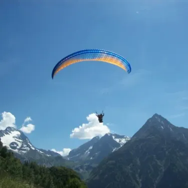 Parapente bleu-jaune volant au-dessus d'une vallée montagneuse sous un ciel bleu.