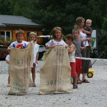Enfants à Camping Le Colporteur portant des sacs en toile sur un terrain gravillonné.