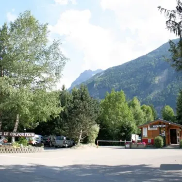 Camping Le Colporteur : entrée du camping avec un bâtiment de réception, des arbres et des montagnes en arrière-plan.