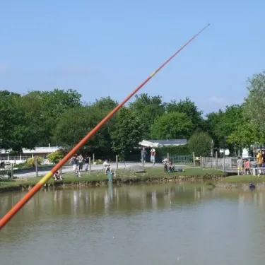 Pêche au lac avec un long bâton de pêche en premier plan, des visiteurs autour d'une zone de pêche au bord d'un étang.