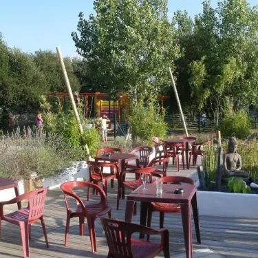 Camping La Baie Dorée : terrasse avec tables et chaises rouges, jardin verdoyant et statue de Bouddha.