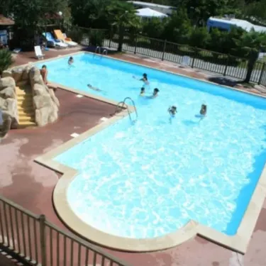 Piscine avec toboggan rocheux et enfants nageant, entourée d'arbres et de chaises longues.